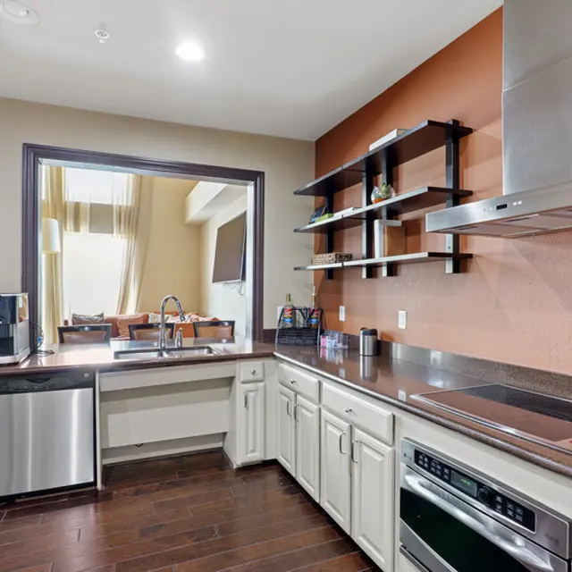 A modern kitchen with a dark wood floor, light-colored cabinets, and stainless steel appliances. A large window allows natural light to flood the space, complementing the warm colored walls.