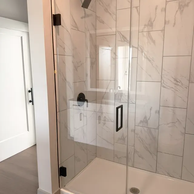 A modern walk-in shower with glass doors featuring marble tiles and a black shower head. The shower is enclosed, and there is a glimpse of a bathroom vanity in the reflection of the glass. The floor is wooden, and a white door is visible in the background.