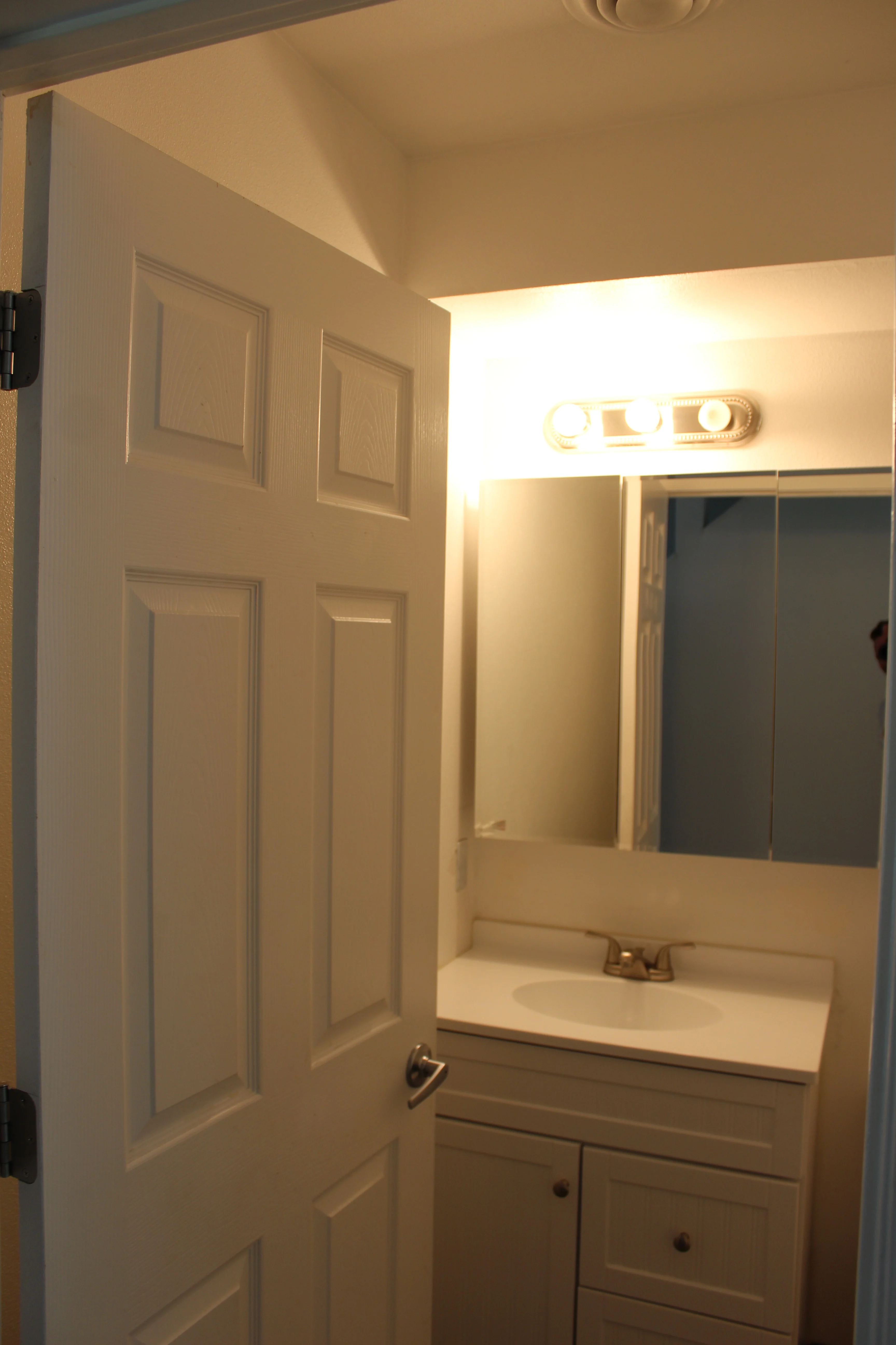 Interior view of a bathroom featuring a white vanity, mirror, and overhead lighting with an open door.