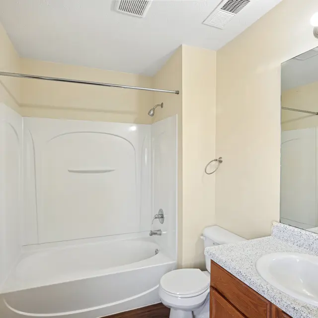 Modern Bathroom Interior A small bathroom featuring a bathtub with a shower, a toilet, and a vanity with a sink and mirror. The walls are painted in a light beige color, and there is a wood floor. A towel rack is mounted on the wall near the shower.
