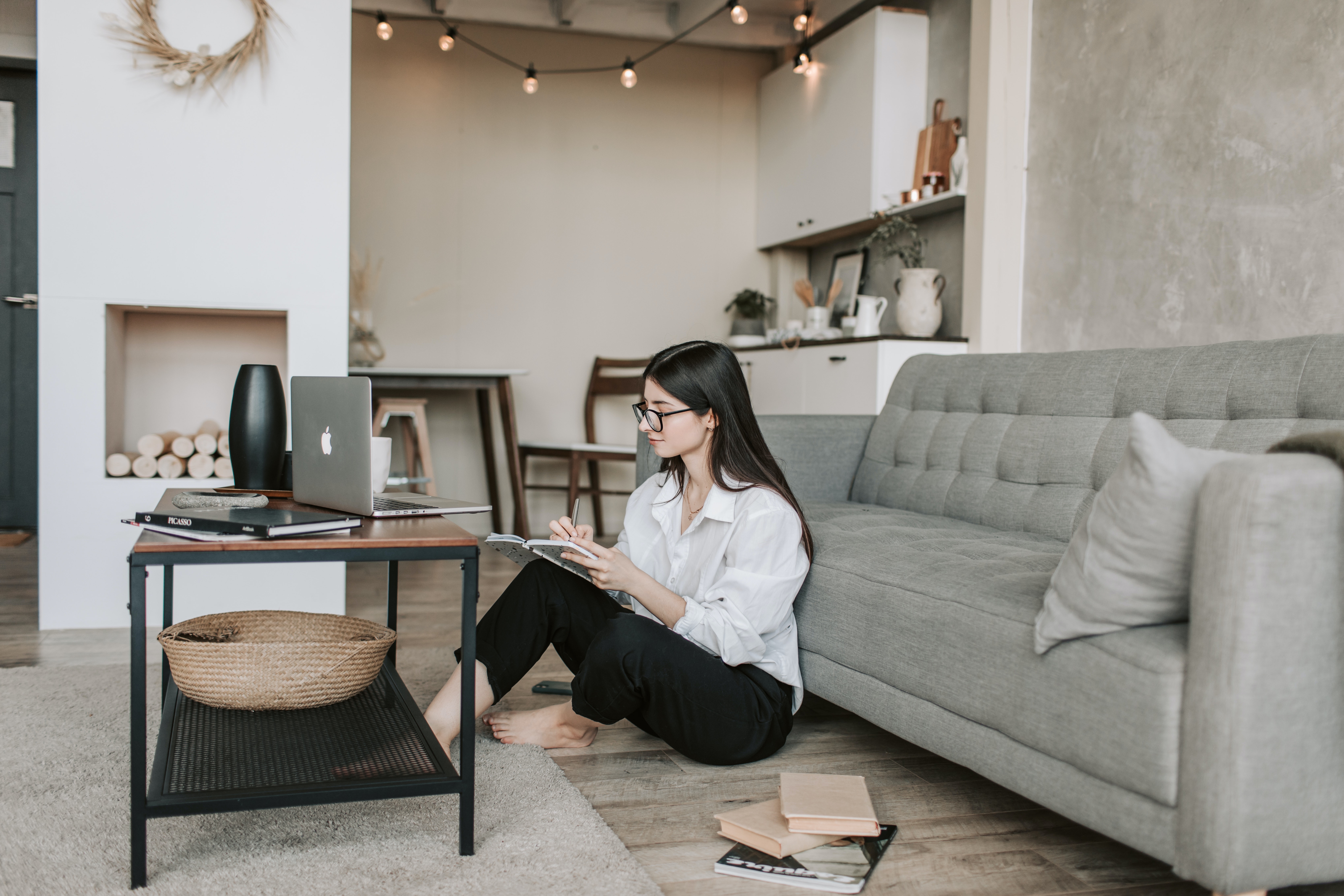 Chic Home Workspace A woman sitting on the floor in a modern living room, working on a notebook while seated next to a coffee table with a laptop and a basket. She is wearing a white shirt and black pants, with long hair and glasses, in a minimalist styled room.