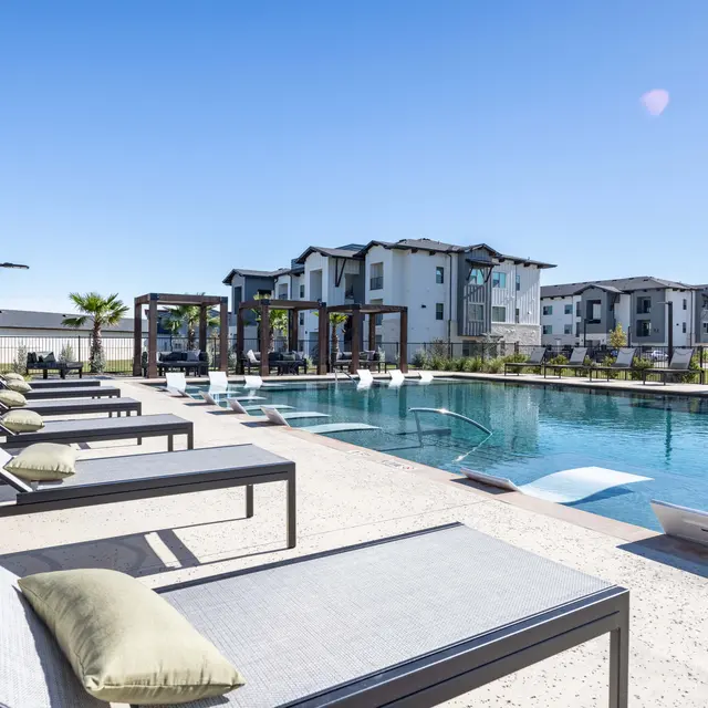 A modern apartment pool area featuring lounge chairs, a swimming pool, and contemporary architecture in the background.