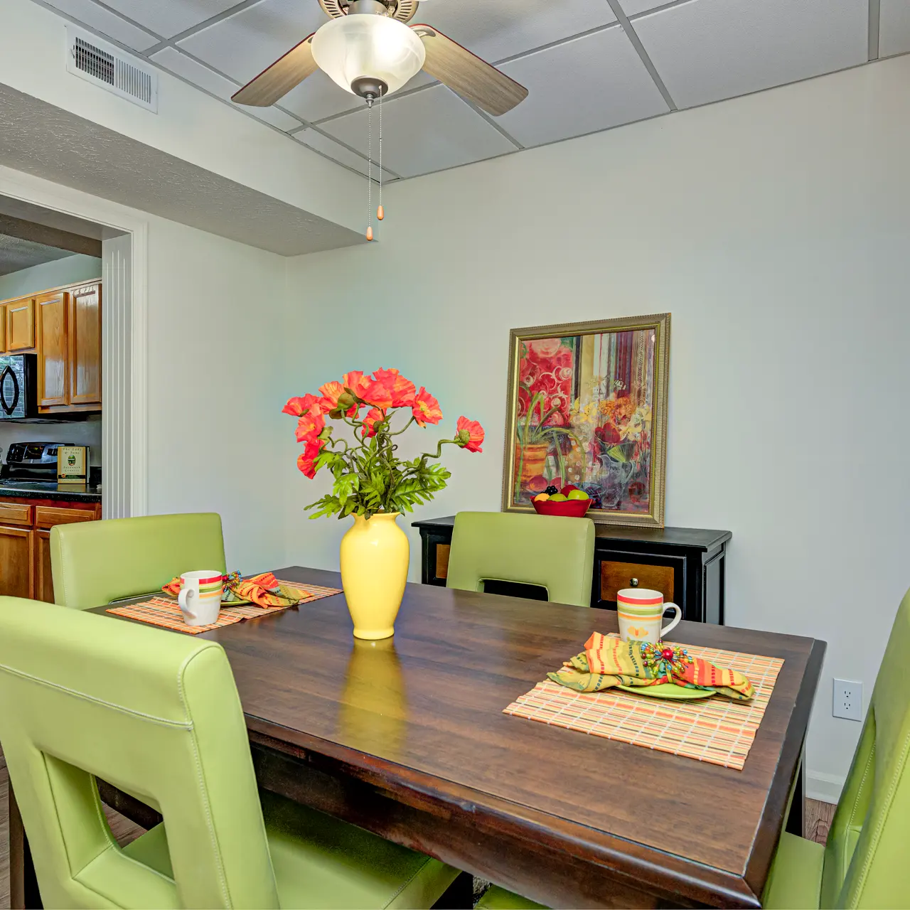 Modern Dining Area A modern dining area featuring a rectangular wooden table with green upholstered chairs. A small vase with pink flowers sits on the table along with two cups. The room has a warm atmosphere, visible kitchen in the background, and light-colored walls.