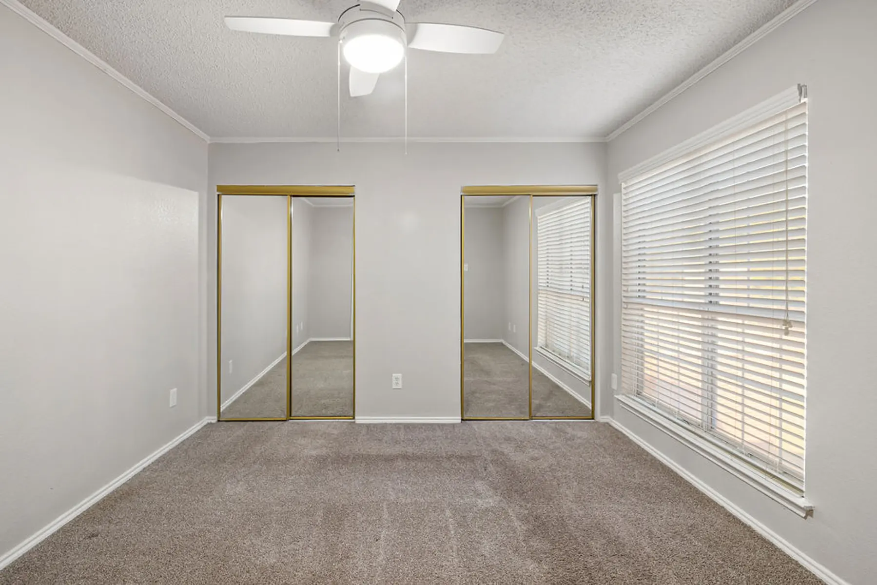 Spacious Empty Room with Mirrored Closets A spacious empty room featuring carpeted flooring and two mirrored sliding door closets. Large windows provide natural light, illuminating the pale gray walls and ceiling. A ceiling fan is present above, enhancing the airy feel of the space.