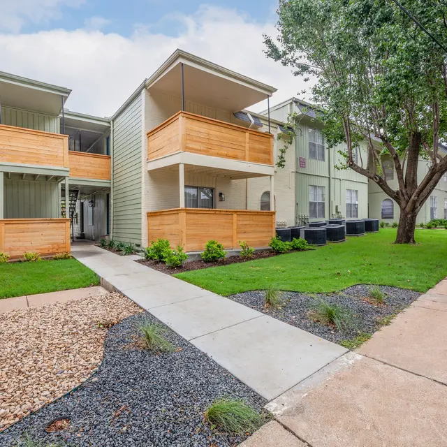 The Hills - A view of an apartment complex featuring two buildings. The first building has wooden balconies and greenery around it. The walkway is lined with gravel and grass, leading to the second building on the right. Cloudy sky is visible above.