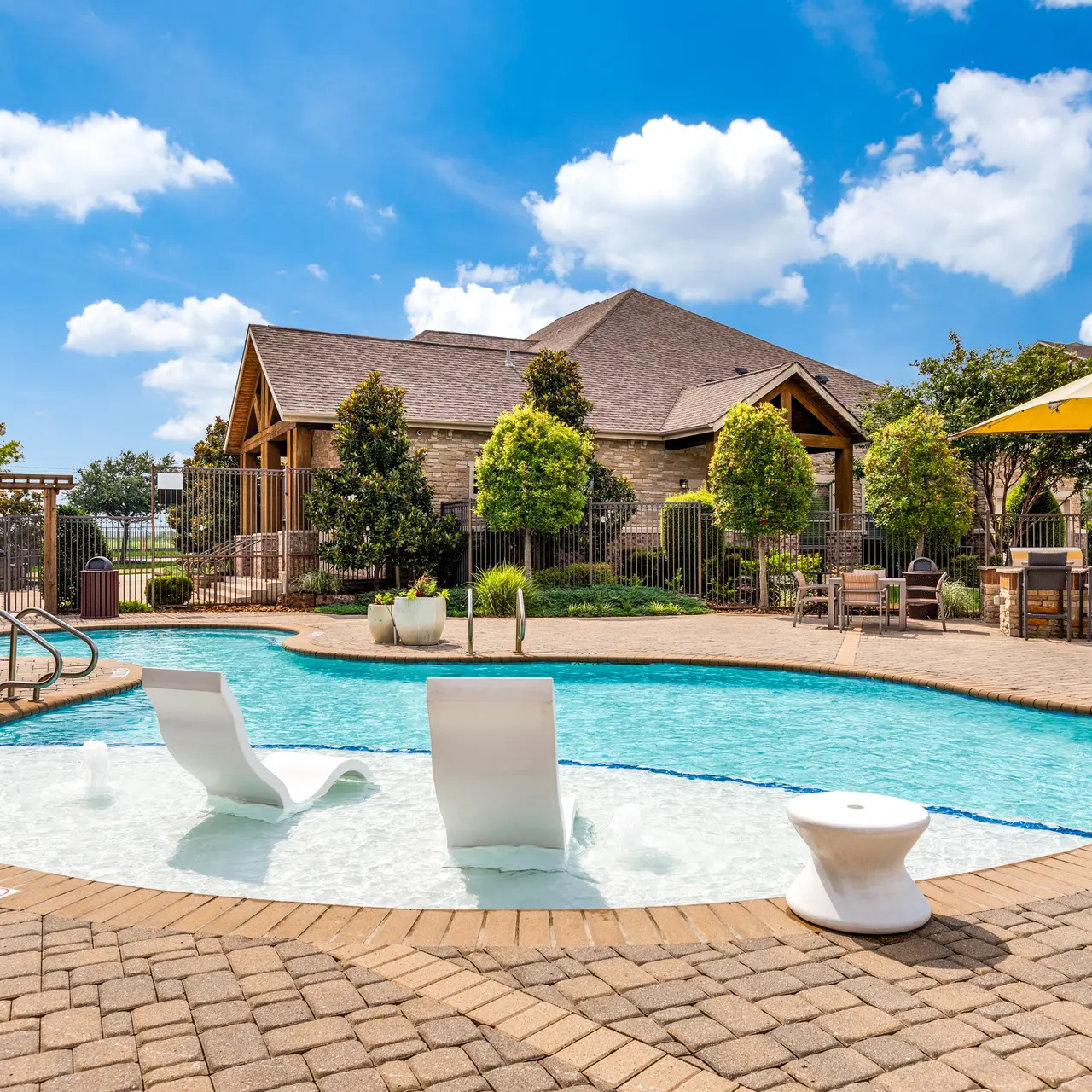 A serene outdoor pool area featuring a curved pool with a shallow section, surrounded by lounge chairs, large umbrellas, and green landscaping. A clubhouse is visible in the background.