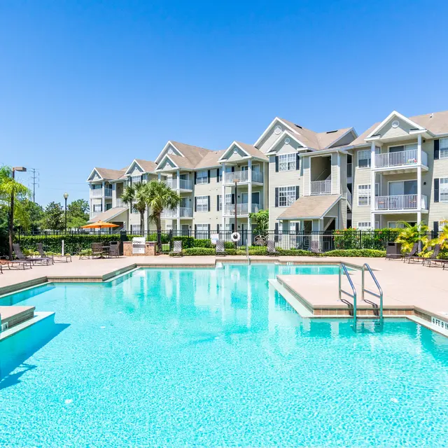 A sparkling blue swimming pool surrounded by palm trees and lounge chairs, with modern apartment buildings in the background under a clear blue sky.