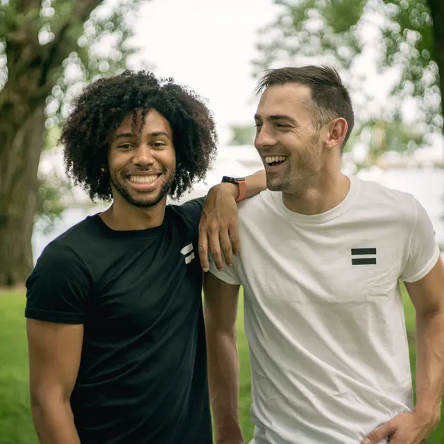 Friends Laughing in Nature Two friends posing outdoors, one in a black t-shirt and the other in a white t-shirt, both smiling and looking happy.