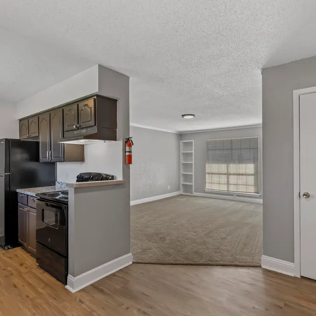 Open concept kitchen and living room area featuring a modern design with wooden flooring. The kitchen includes black appliances and dark cabinetry, while the living room has a large window with natural light.