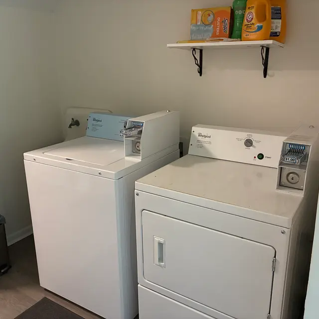 A laundry room featuring a white washing machine and dryer side by side. Above them, a shelf holds laundry detergent and fabric softener. A trash bin is visible in the corner.