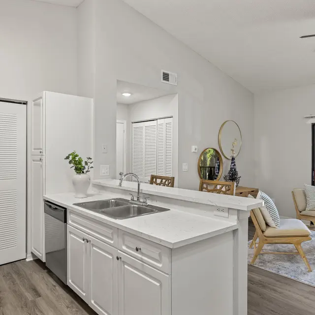 A modern kitchen with white cabinets and a dishwasher, looking towards a cozy living room area with a view of greenery through the large window.