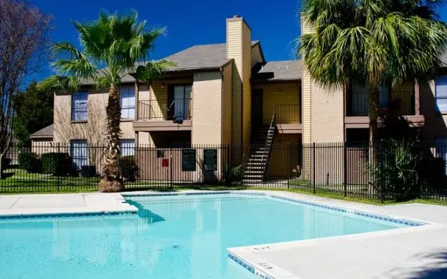 A serene apartment complex featuring a swimming pool surrounded by palm trees, with a two-story building in the background and clear blue skies above.