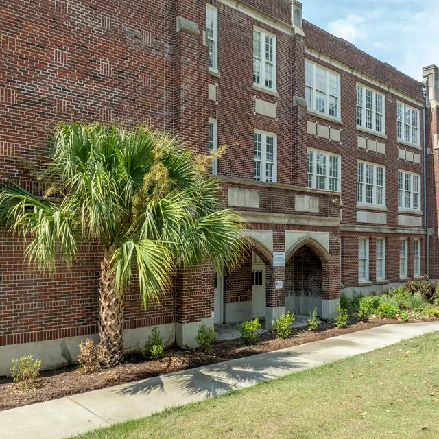 A historic brick building with large windows and an archway entrance, flanked by palm trees and landscaped greenery.