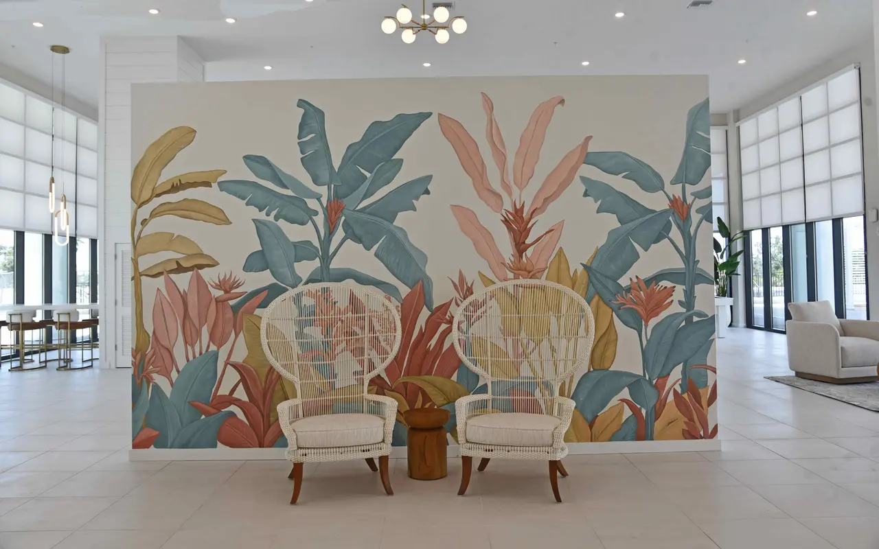 A modern lobby featuring a large floral mural with tropical plants, two white chairs, and a small wooden side table. The space has bright lighting and large windows.