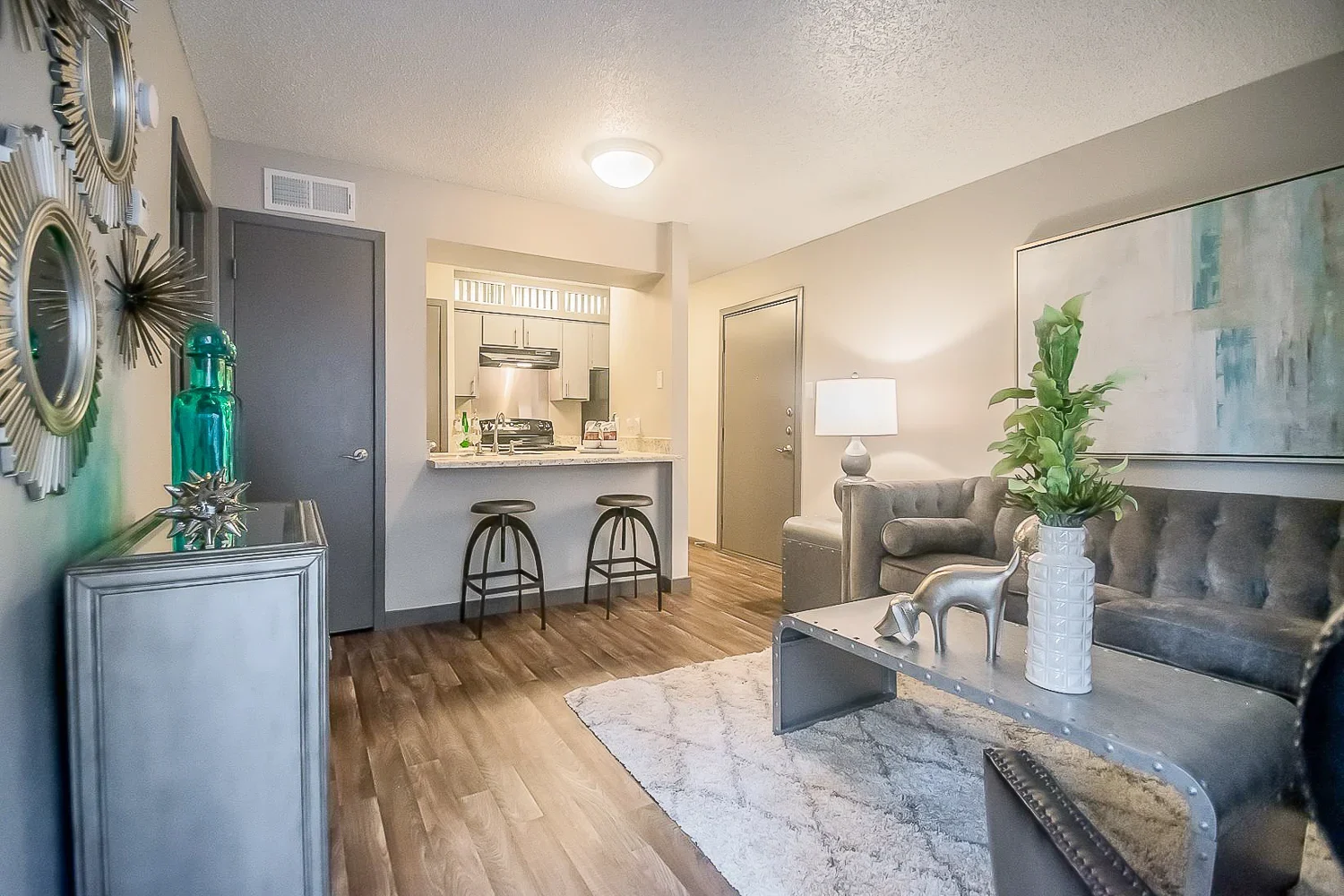 A modern living room featuring a gray tufted sofa, a light-colored area rug, and a stylish coffee table. There is a kitchen area visible in the background with bar stools and a countertop. Decorative plants and wall mirrors add elegance to the space.