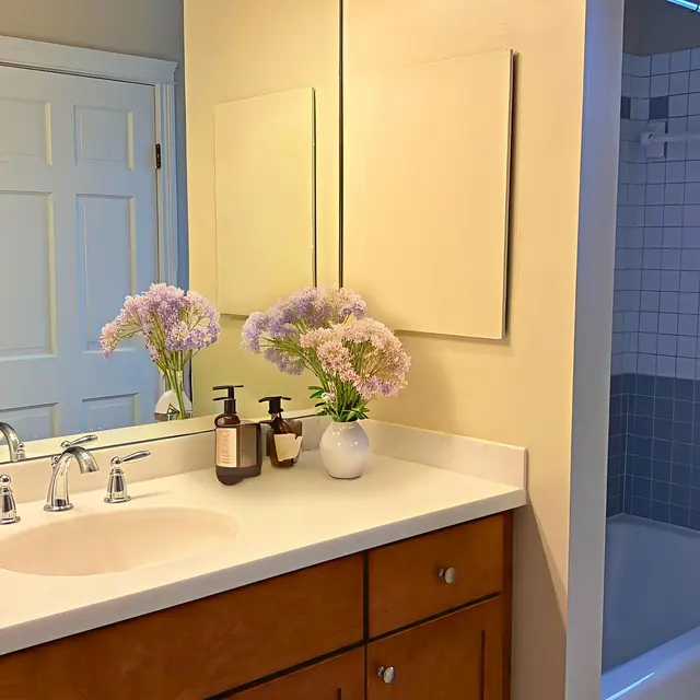 A well-lit bathroom featuring a countertop with a sink and faucet, adorned with flowers in decorative pots. A large mirror reflects the scene, and a bathtub is visible in the background. The decor includes beige walls and wood cabinetry.