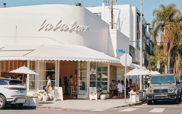 A trendy café with outdoor seating under white umbrellas is seen at the corner of a street, surrounded by palm trees. Vehicles are parked along the street, and people are enjoying their time at the café.