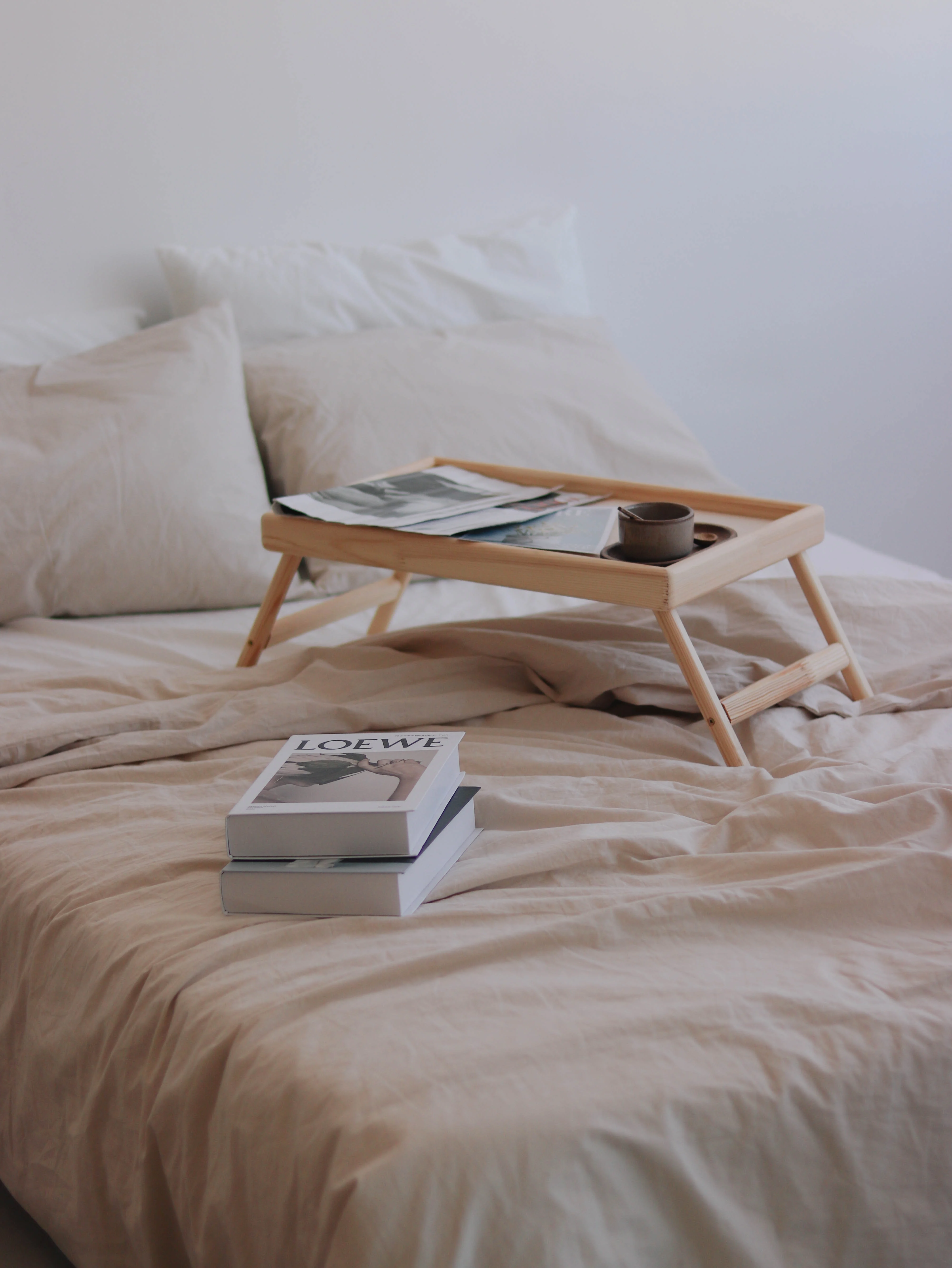 Cozy Bedroom Setup with Tray and Books A cozy bedroom scene featuring a bed with beige linens, a tray holding magazines and a cup, along with a stack of books.
