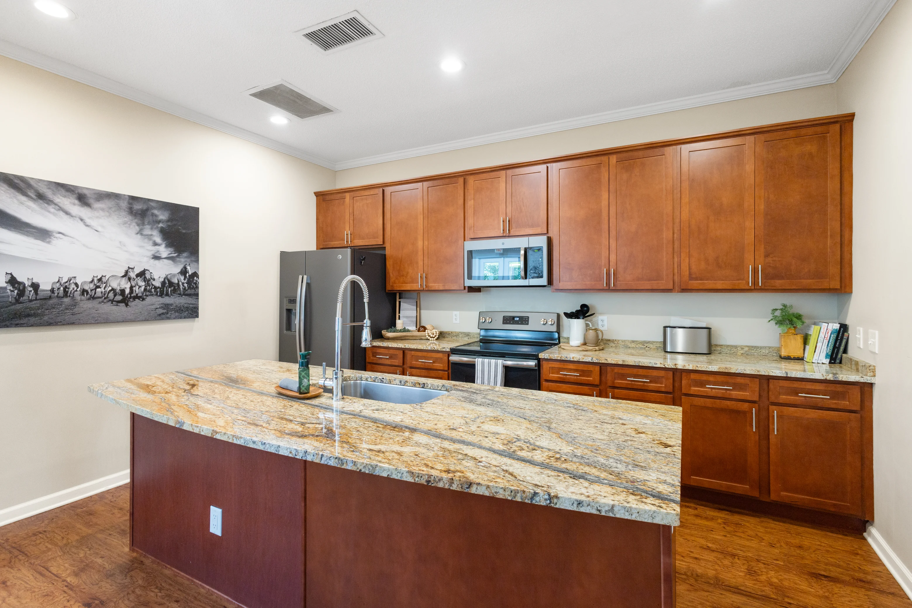 A spacious modern kitchen with wood cabinetry and a large granite island, featuring stainless steel appliances and a wall photo of horses.