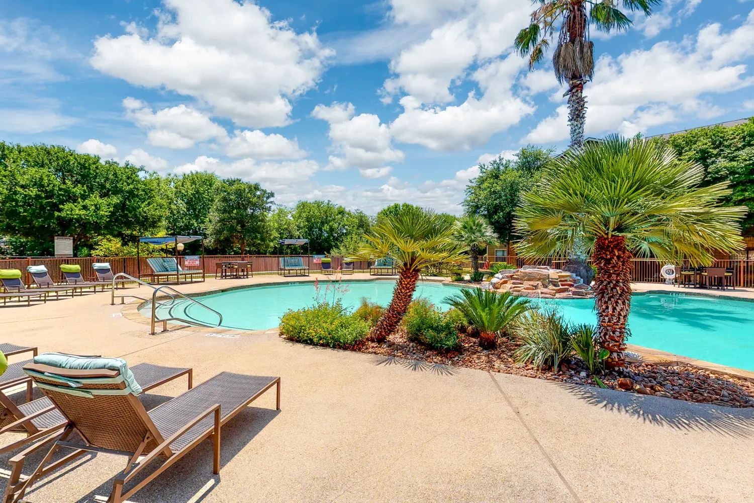 Swimming Pool Area A spacious swimming pool area surrounded by palm trees and lounge chairs under a blue sky with fluffy clouds.