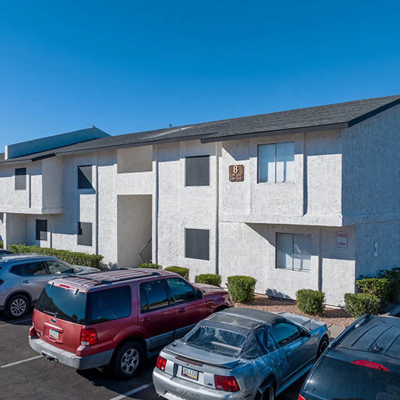 Exterior view of a two-story apartment building with a grey roof and white walls, surrounded by parked cars in a parking lot.