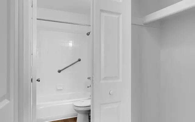 A view of a bathroom from a doorway, featuring a white tub, toilet, and wooden flooring. The bathroom walls are white, and there is a grab bar installed near the tub.