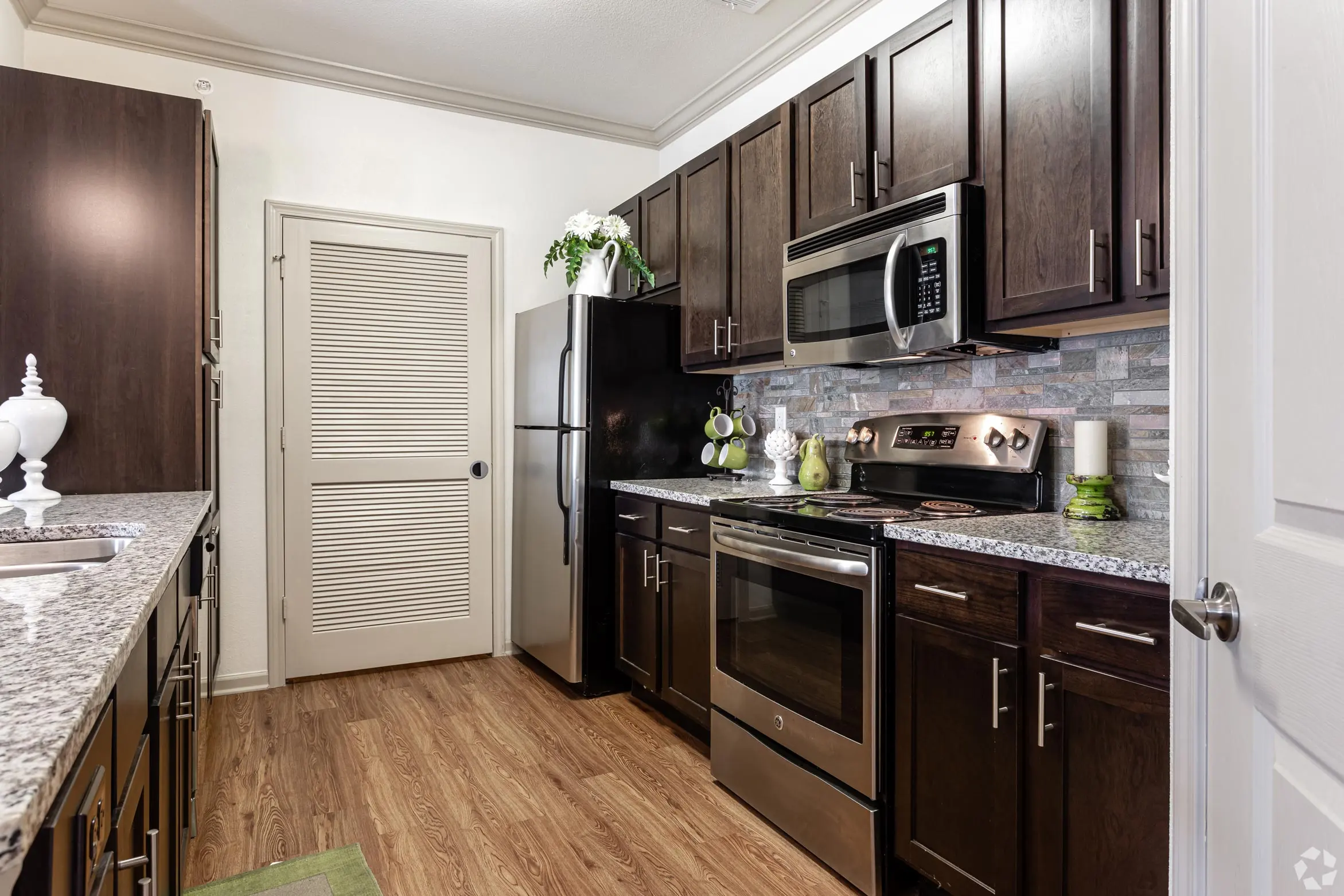 Modern Kitchen Design A modern kitchen featuring dark wooden cabinets, stainless steel appliances, and granite countertops. The room has an organized layout with a door leading to another area and decorative plants on display.