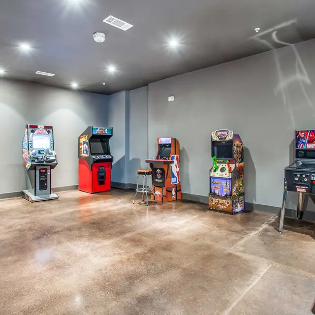 An arcade room featuring several vintage arcade game machines and a flat-screen television, with polished concrete flooring and gray walls.
