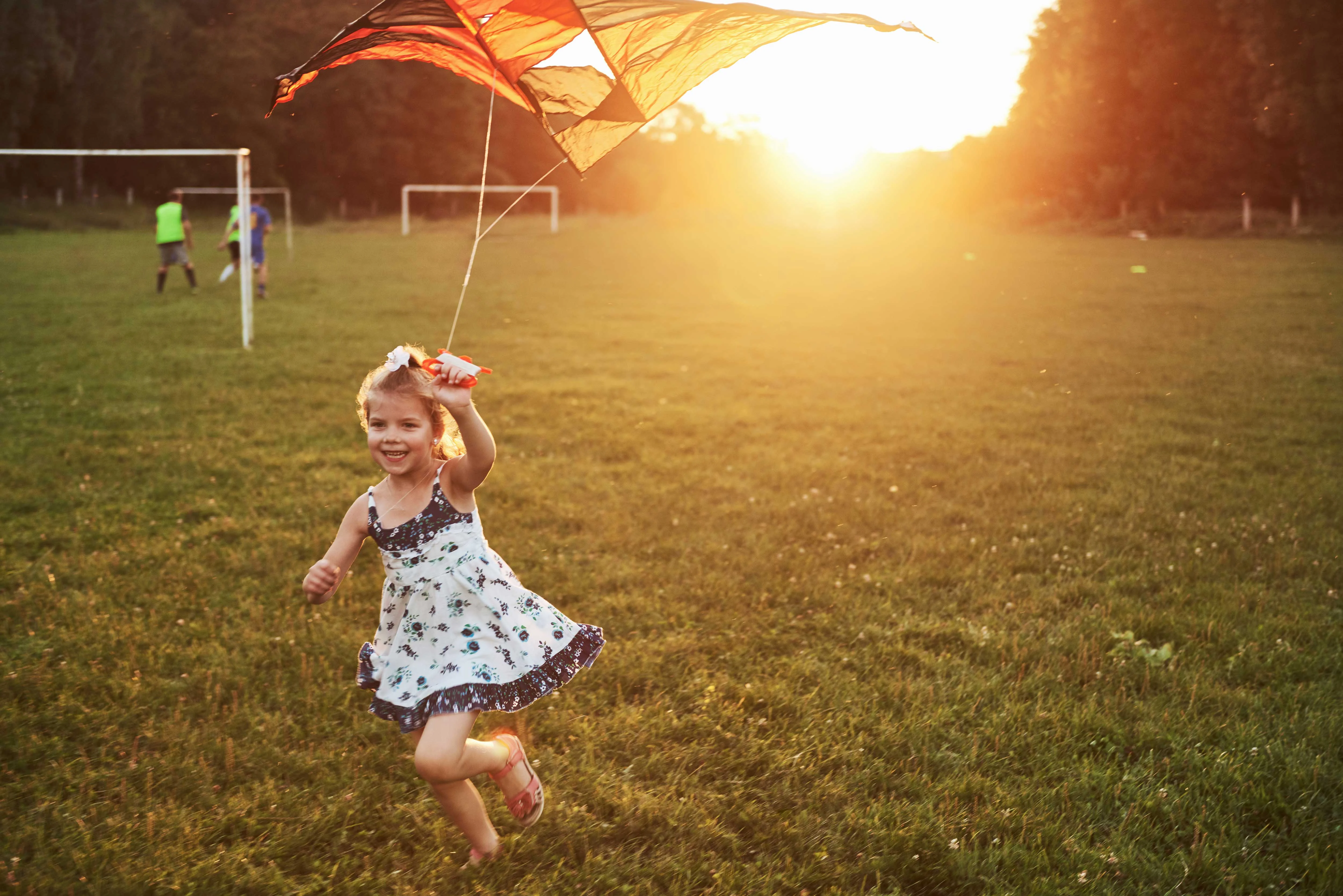 Girl Flying a Kite at Sunset A young girl in a floral dress joyfully runs with a kite against a sunset backdrop on a grassy field. In the background, there are soccer goalposts and other individuals playing soccer.