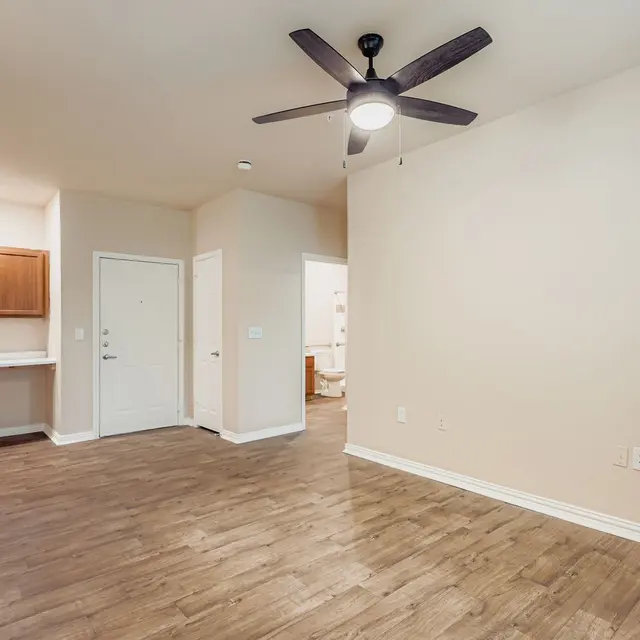 Modern Apartment Interior A spacious, modern apartment interior featuring hardwood flooring, a ceiling fan, and light beige walls. On the left, there is a kitchenette with wooden cabinets. The entryway is seen at the center, leading to another room in the background.
