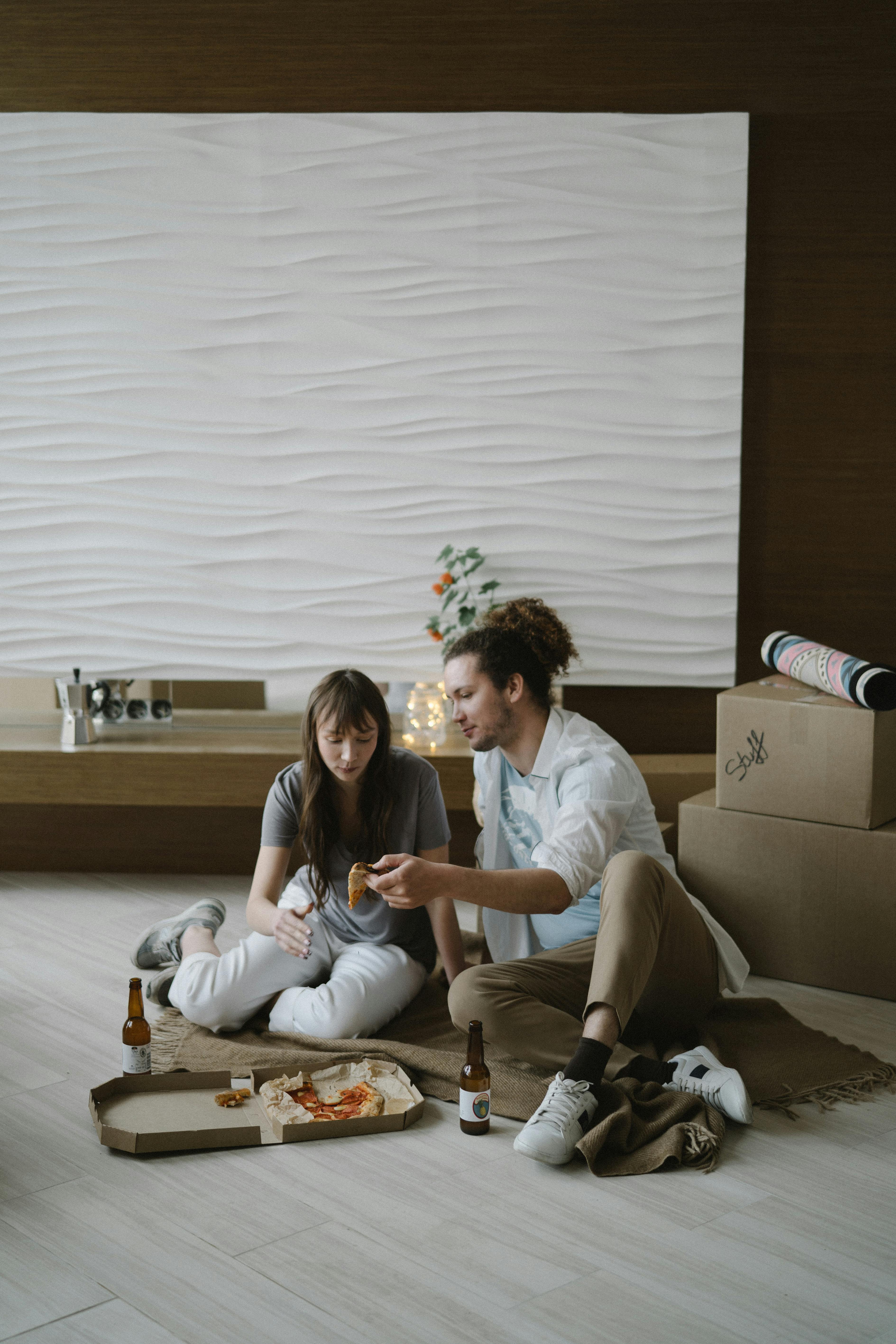 A couple sitting on a soft rug, sharing a pizza and drinks in a cozy living space with a modern design.