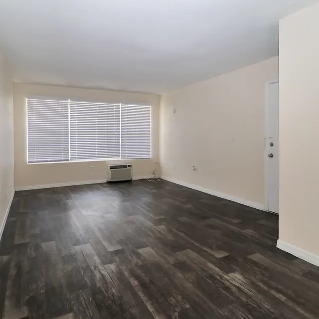 A spacious, empty room with light-colored walls and dark wooden flooring. There are large windows with blinds allowing natural light to enter, and an air conditioning unit on the wall. A door leads to another area.
