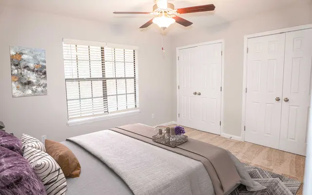 Cozy bedroom with a ceiling fan, two windows, a neutral color scheme, a neatly made bed, and a couple of closet doors.