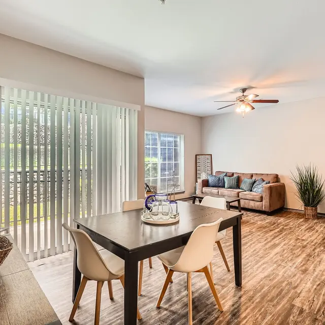 A modern apartment living space showcasing a dining area with a dark table and chairs, a cozy living area with a brown sofa, and large windows allowing natural light. The flooring is wooden, and there's a plant in the corner.