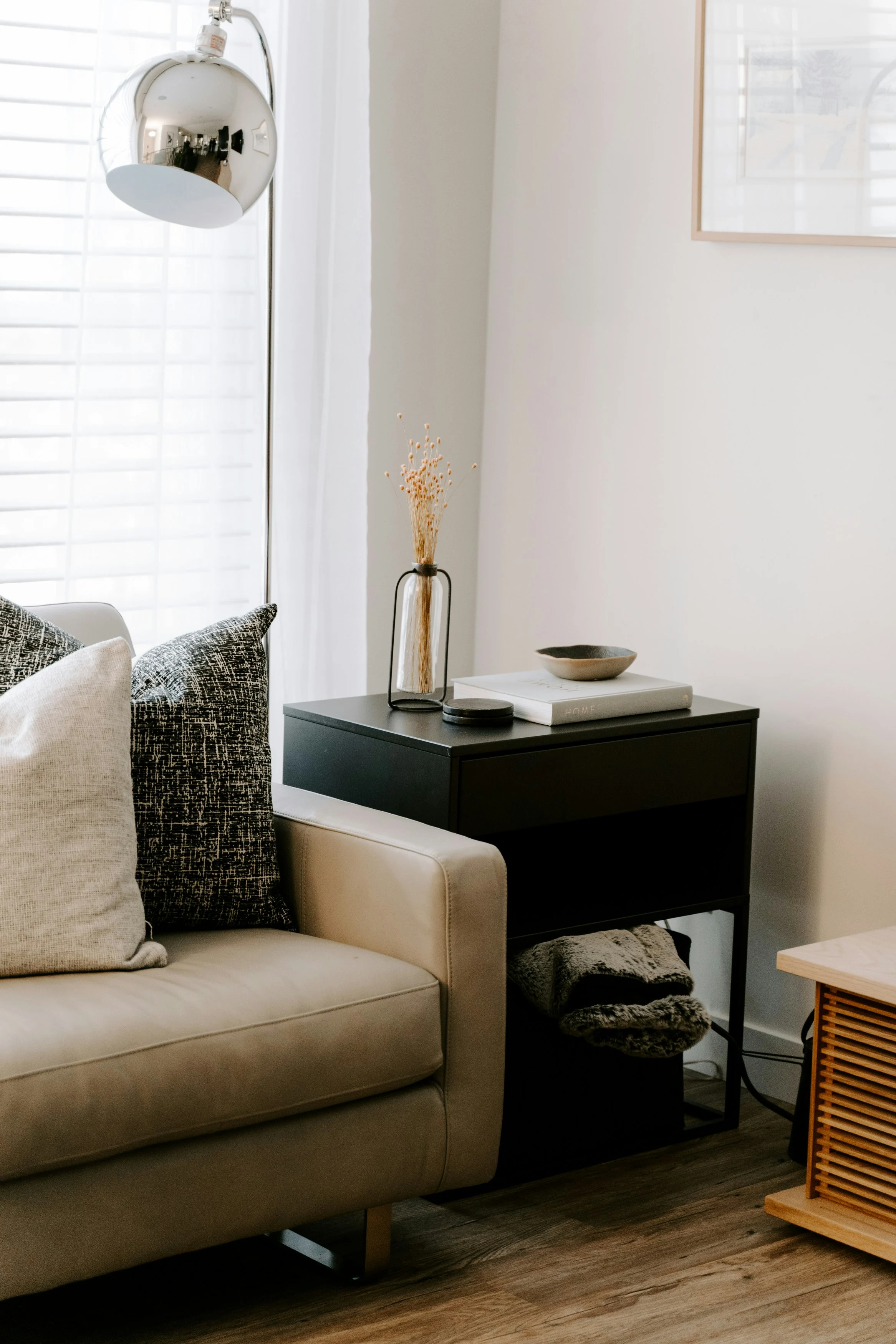A cozy corner of a living room featuring a light beige sofa with decorative pillows, a sleek black nightstand holding decorative items, and a stylish silver lamp.