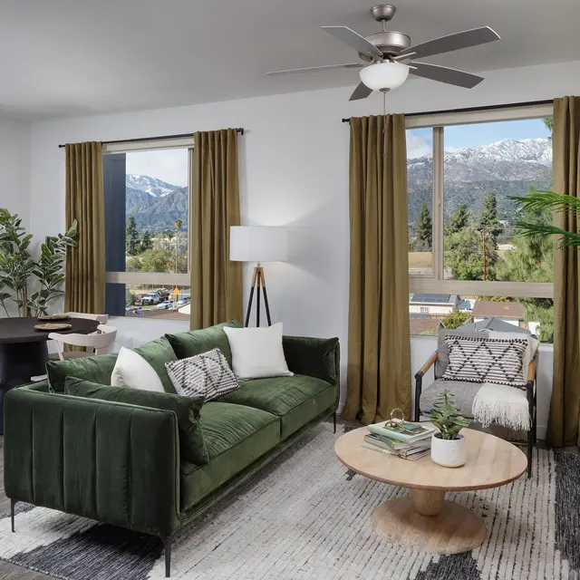 A modern living room featuring a green velvet sofa, a round wooden coffee table, and large windows with mountain views. The room includes a dining area with a small table and chairs, plants, and decorative elements.