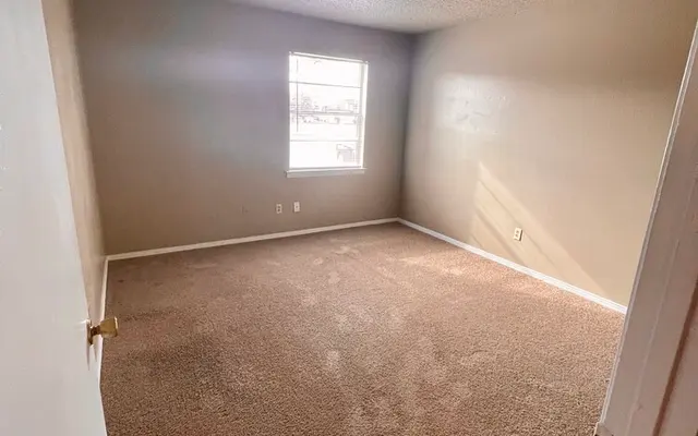 Empty Room Interior A vacant room with light brown carpet, beige walls, and a single window allowing natural light.