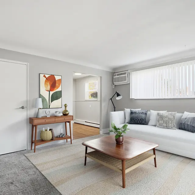 A cozy modern living room featuring a white couch, wooden coffee table, and decorative items. The walls are painted light gray with a colorful flower painting on display. Large windows with vertical blinds allow natural light to fill the space, and there is a wooden accent table beside the couch.