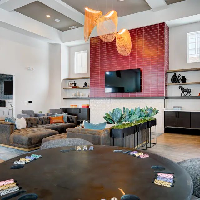 A modern living room with a large sectional sofa, a circular table with colorful chips, and a red tiled wall featuring a mounted TV. Decorative plants and shelves with various items are also present.