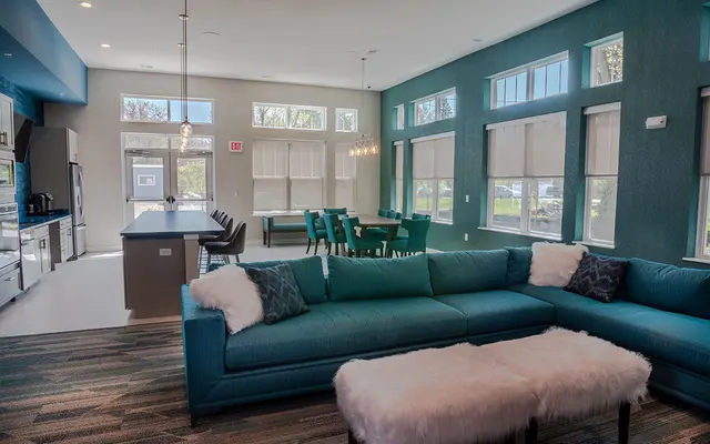 Modern living room with a large blue sofa, white ottoman, teal chairs, and kitchen in the background.