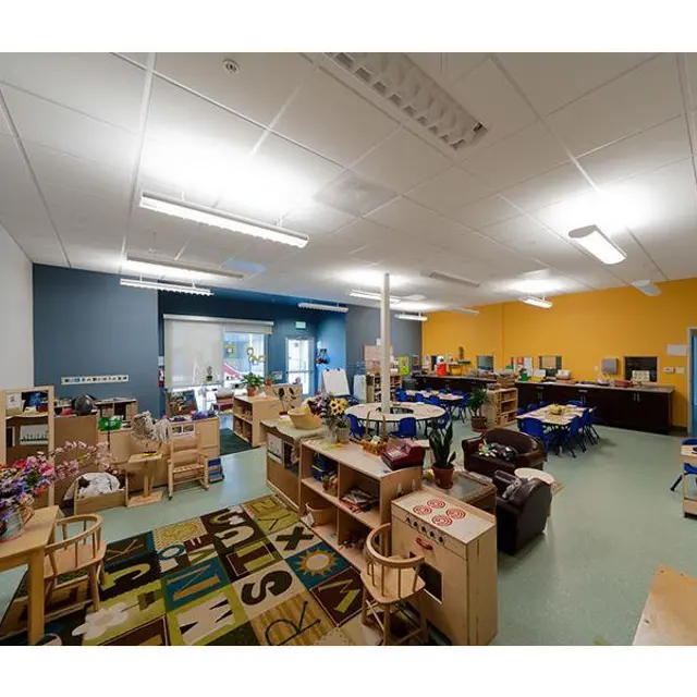 A brightly colored classroom filled with wooden furniture, educational materials, and a colorful rug. There are plants, manipulatives, and a cozy seating area in the space.