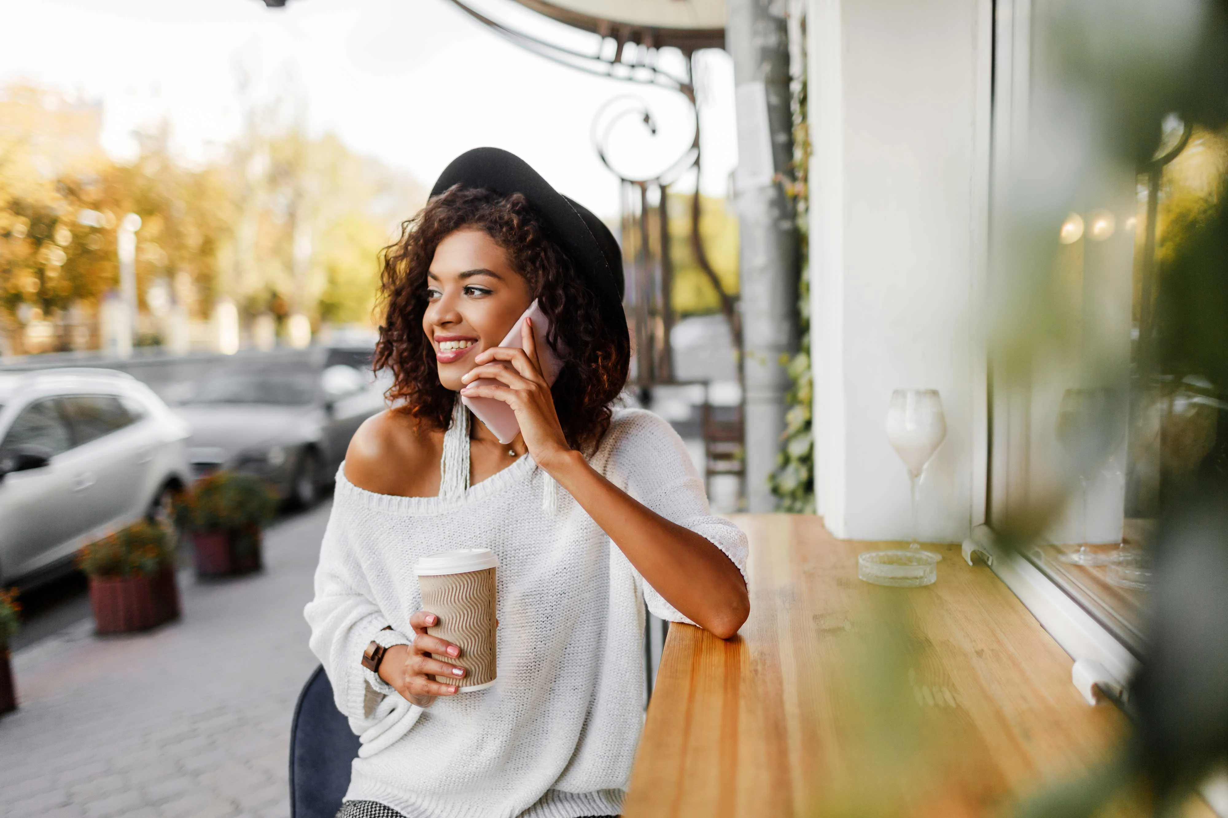 Relaxing at a Café A smiling woman seated at a wooden table outside a café, talking on her phone and holding a coffee cup. She has curly hair and is wearing a cozy sweater and a wide-brimmed hat.