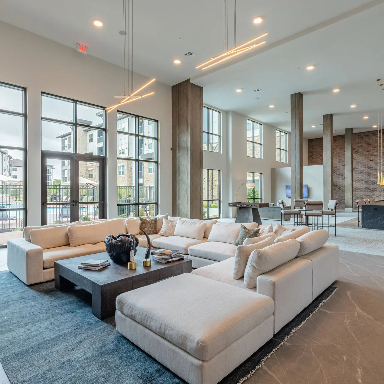 Contemporary Living Room Interior A spacious and modern living room featuring large windows, a comfortable sectional sofa, a coffee table with magazines, and a stylish lighting fixture. The area has a minimalist decor with neutral colors, large floor tiles, and a view of an outdoor area through the windows.