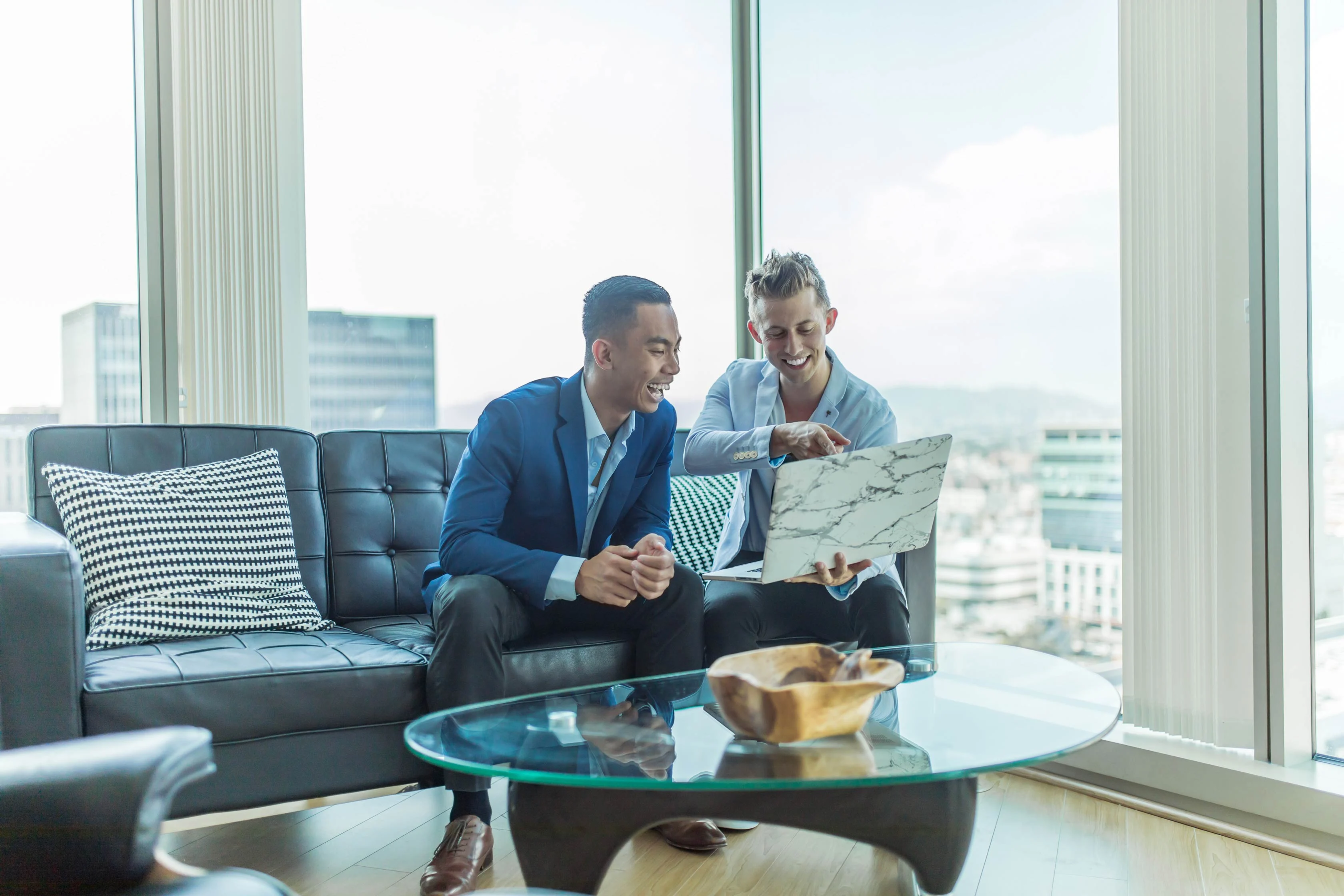 Two men in business attire sitting on a couch, smiling and looking at a laptop in a modern office environment with city views.