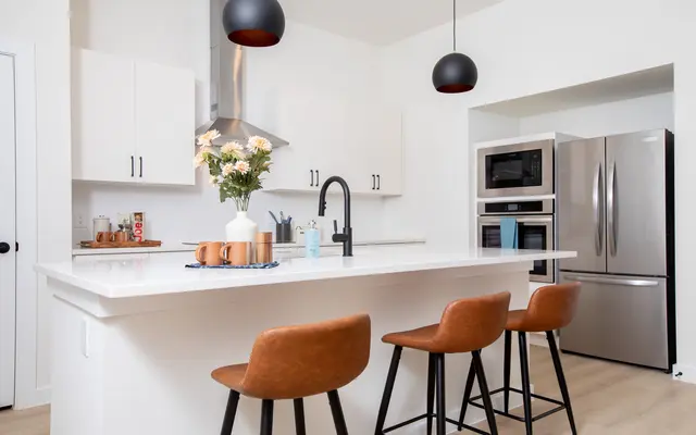 A modern kitchen showcasing a large island with brown bar stools, white cabinets, and stainless steel appliances. A flower vase sits on the counter, and black pendant lights hang above.