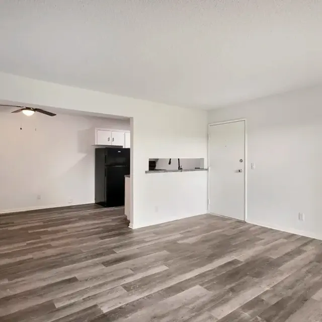A spacious and modern apartment interior with an open layout, featuring a living area with wood-like flooring, white walls, and a door leading outside. There is a partial view of a kitchen area with minimal cabinetry and a ceiling fan above.