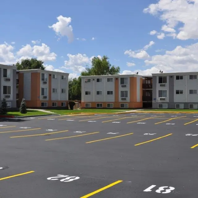 A view of three apartment buildings with a parking lot in the foreground. The buildings are white with orange accents and have multiple windows. A bright blue sky with scattered clouds is visible above.