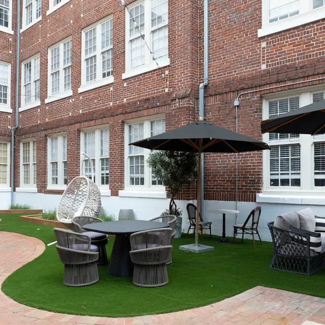 An outdoor seating area featuring a round table surrounded by chairs, a lounge sofa, and umbrellas, set against a backdrop of a brick building with multiple windows.