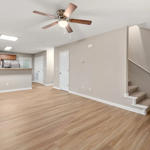 A spacious living area featuring light-colored wooden flooring, a ceiling fan, and a staircase leading to the upper level. The kitchen with wooden cabinets is visible in the background, and there's a doorway leading outside.