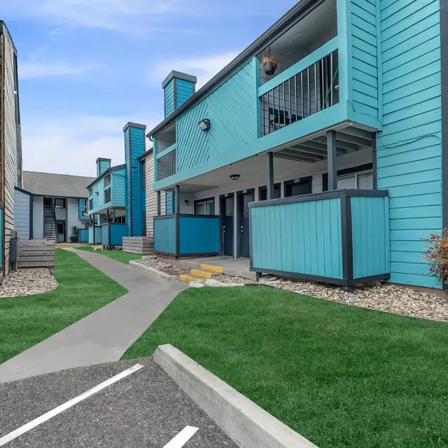 A pathway leading through an apartment complex with blue and grey buildings, lush green grass, and decorative shrubs.